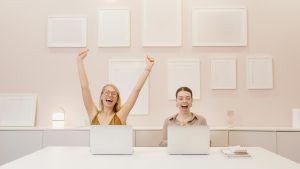 Two women celebrating a success in their modern workspace, working on laptops with blank picture frames on the wall.